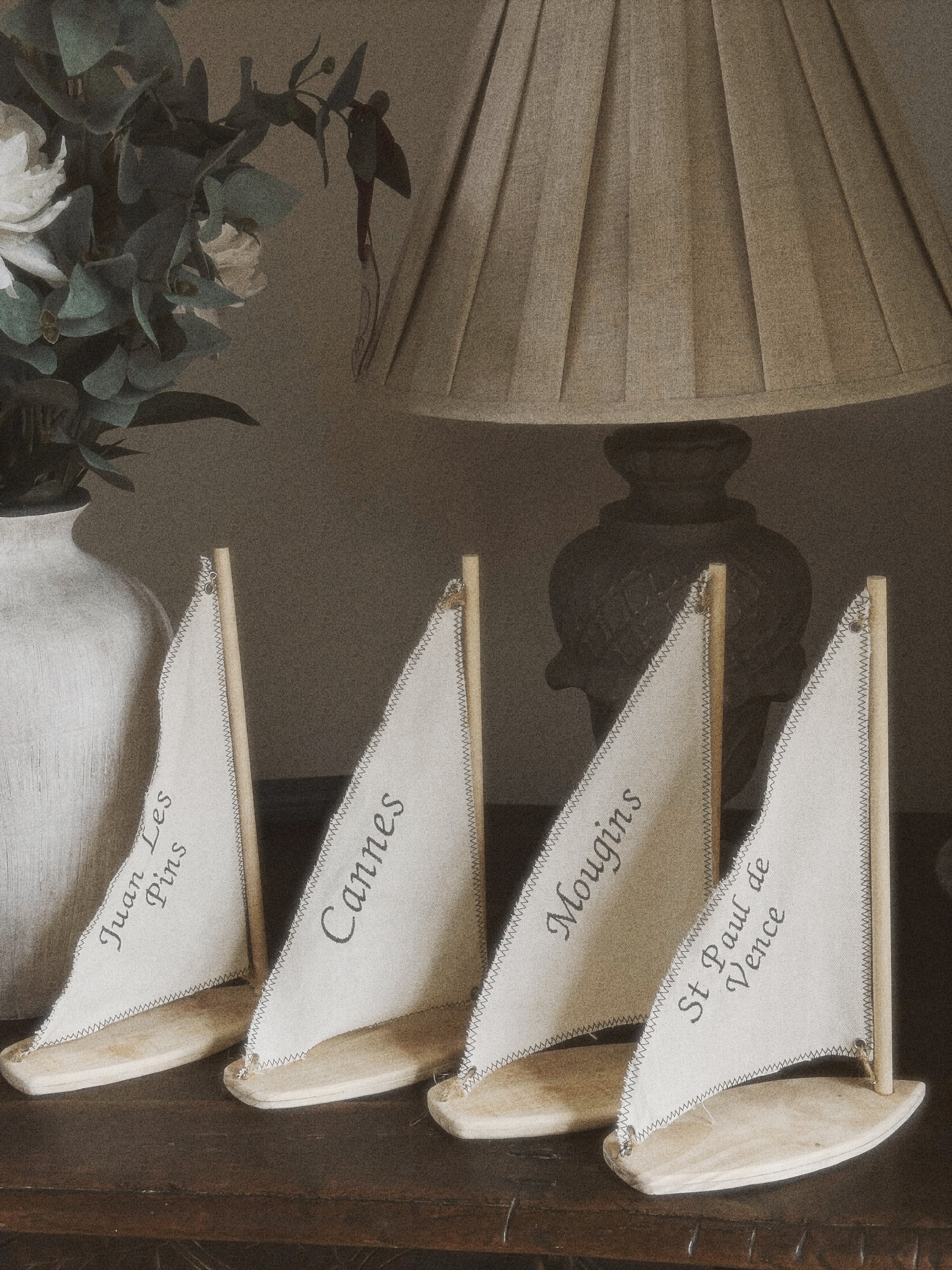 Four small sailboat decorations with cream-colored sails and light wood bases, each printed with a different French coastal city name, arranged on a dark wooden surface next to a lamp and a vase of flowers.