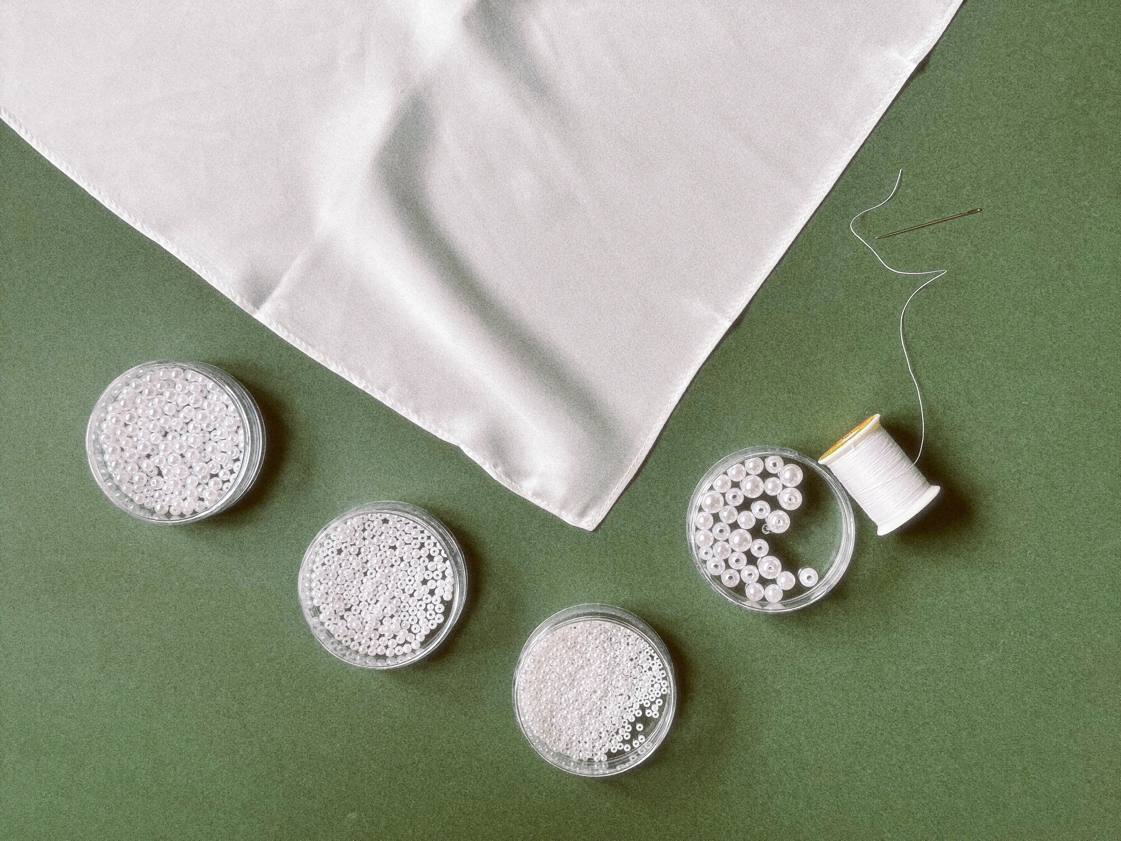 White embroidery floss balls in small clear jars, a spool of white thread, and a folded white fabric on a solid green background.