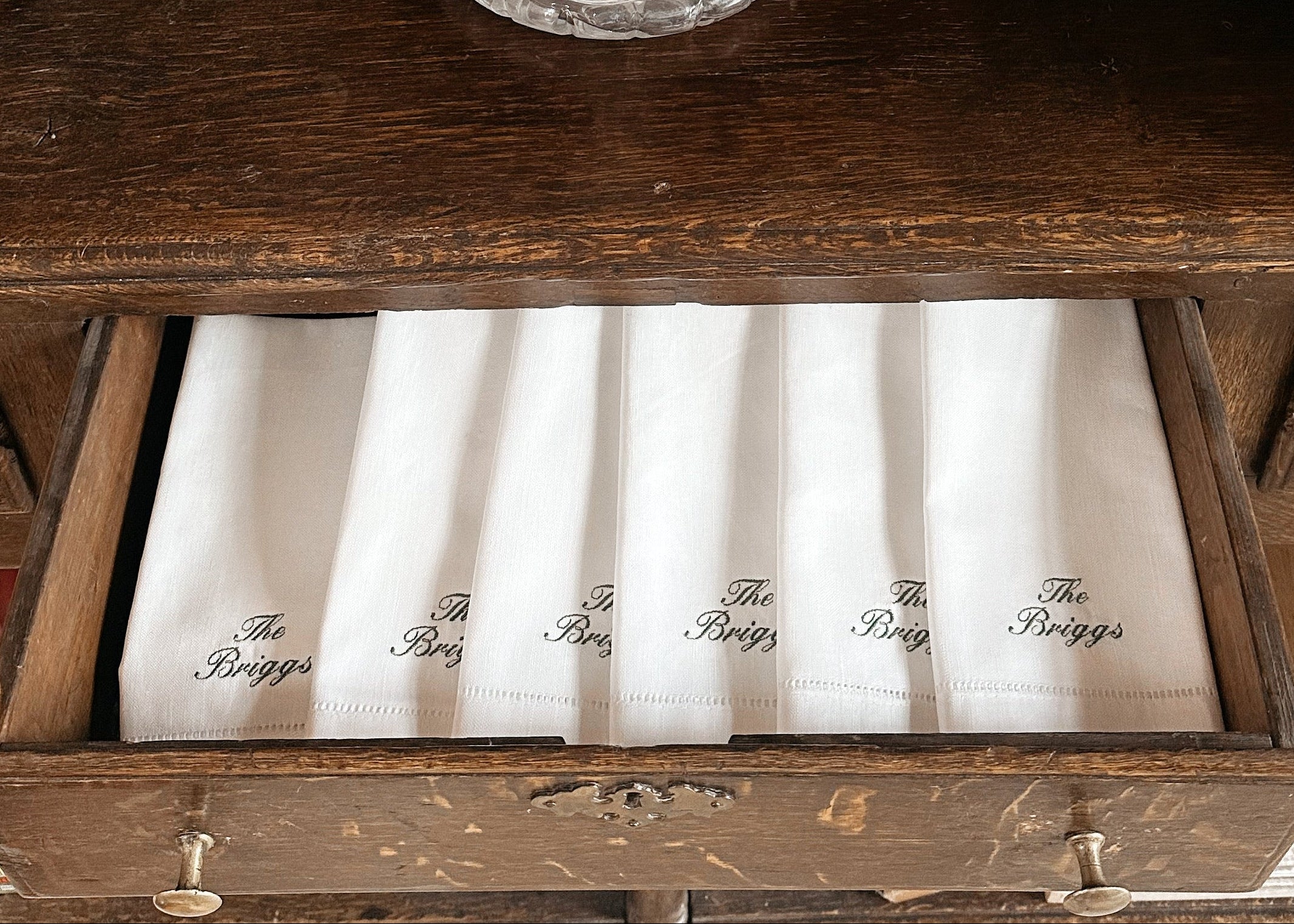 A set of white folded napkins with black script embroidery reading 'The Baggs' arranged in an open wooden drawer, with a clear glass decanter on the shelf above.