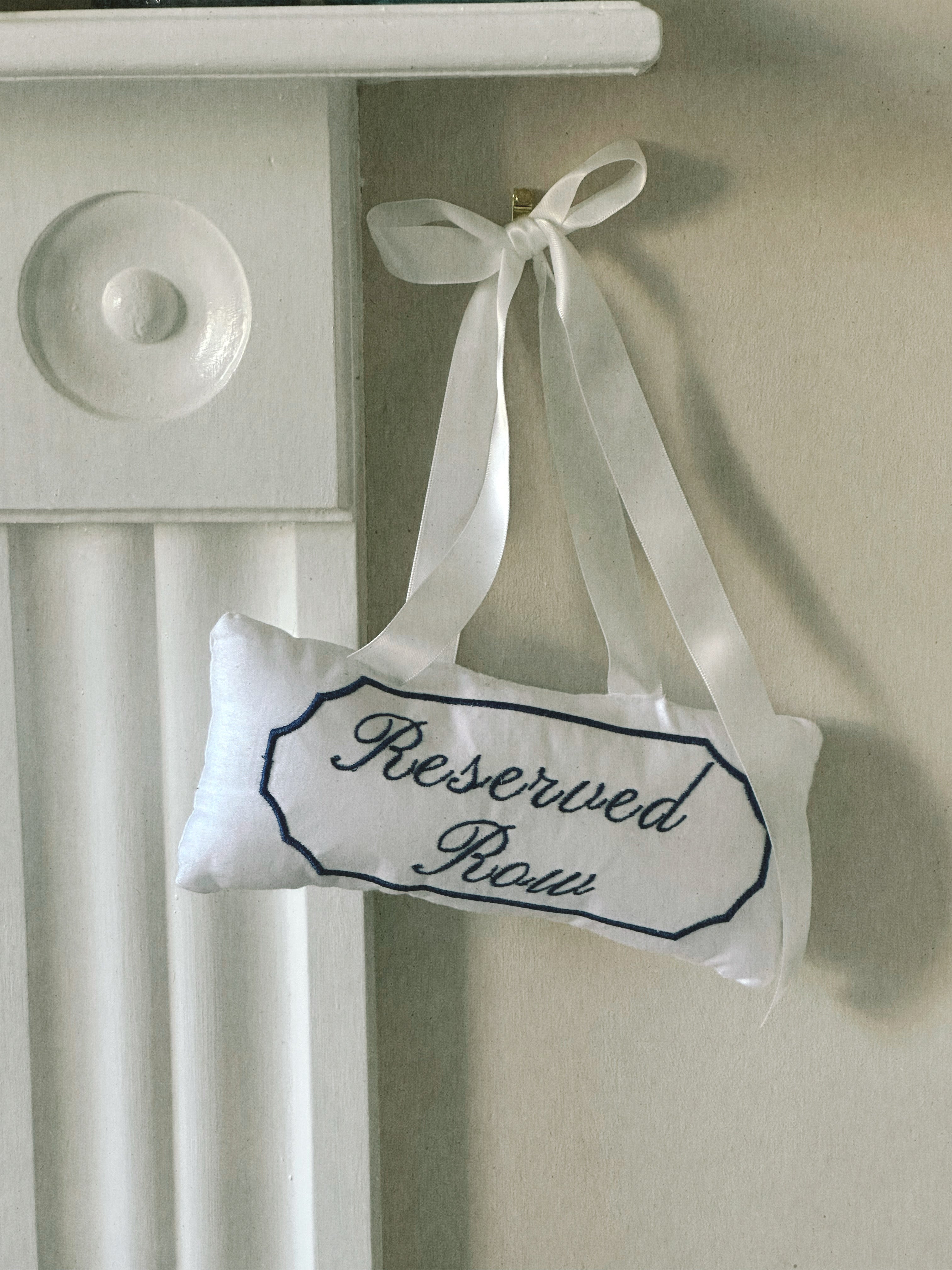 A small white fabric pillow with a navy embroidered border and the words 'Reserved Room' in script, hanging from a white ribbon bow on a beige wall beside a white mantel.