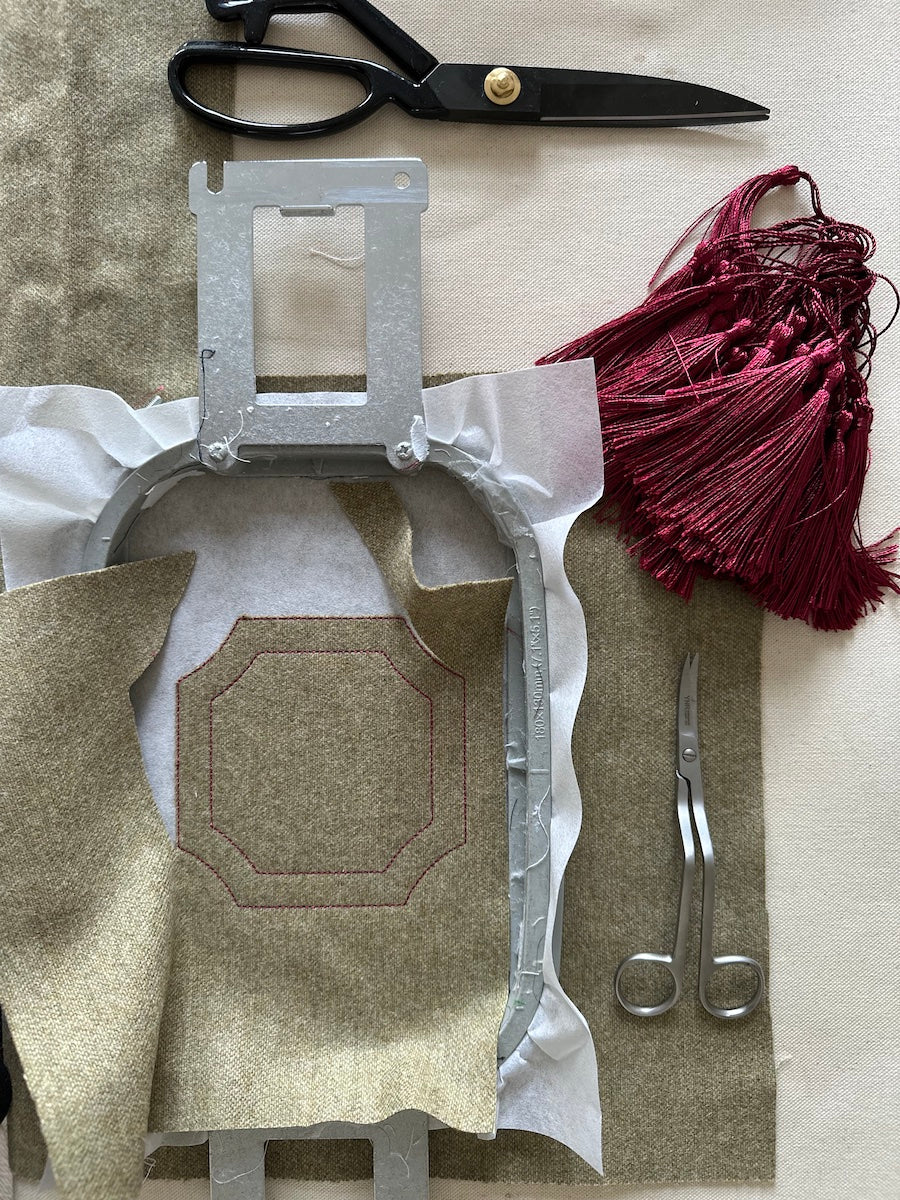 Crafting materials including a metal frame, fabric, scissors, and tassels on a textured surface.